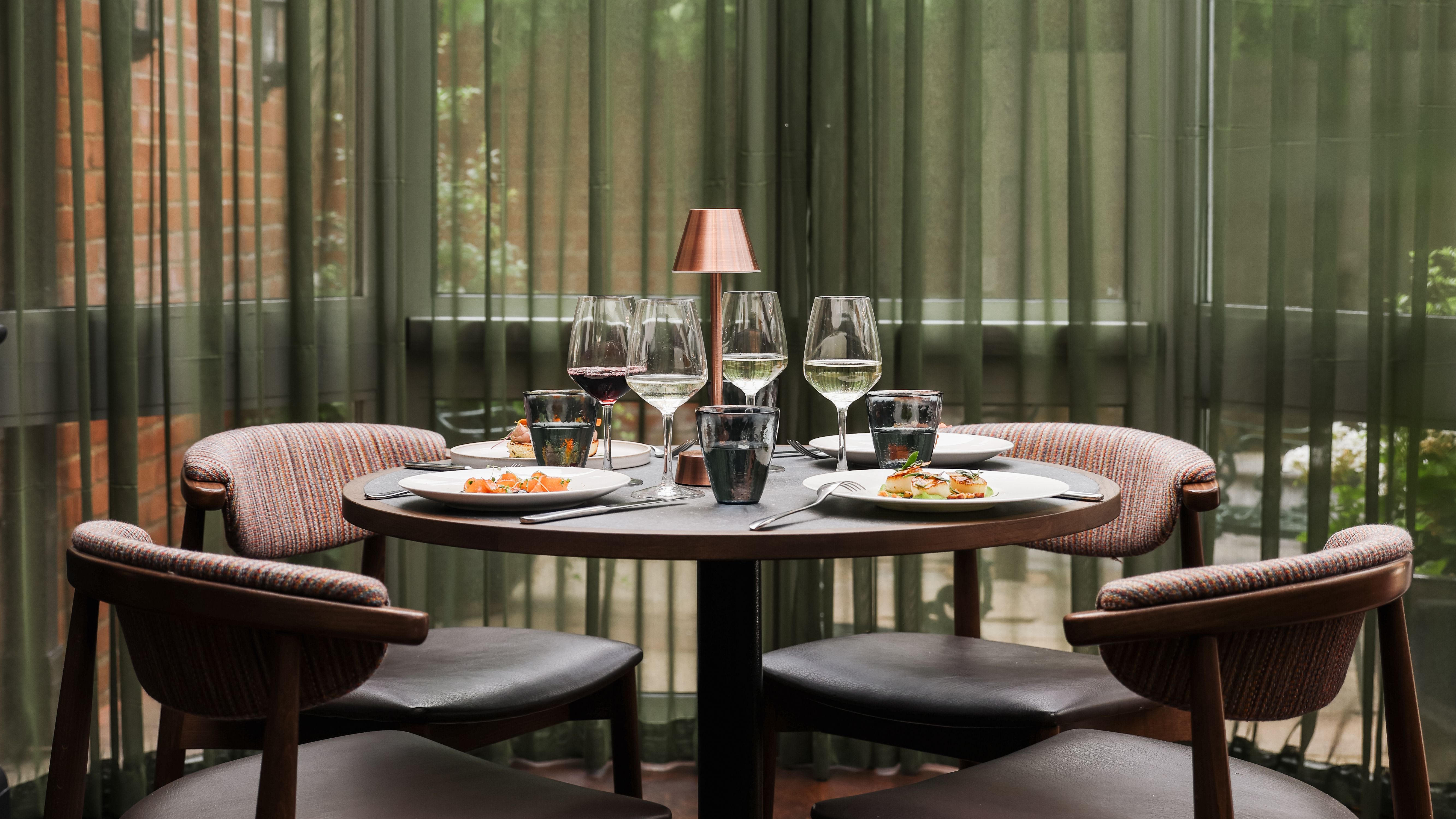 Elegant dining table set for four with wine glasses, plates of food, and a small lamp in a modern restaurant setting.