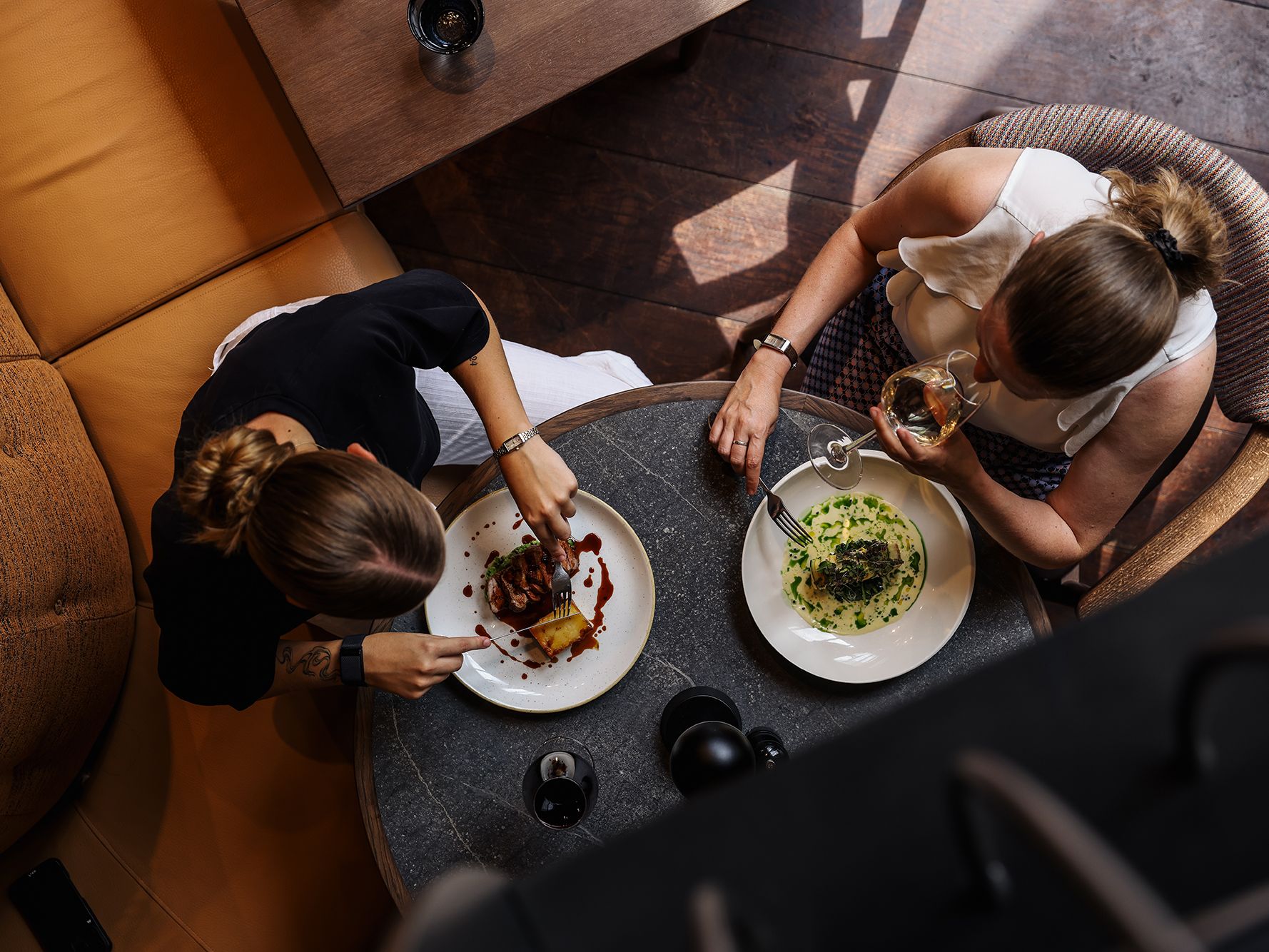 Two women dining at a restaurant table, viewed from above, enjoying plated gourmet meals and drinks.