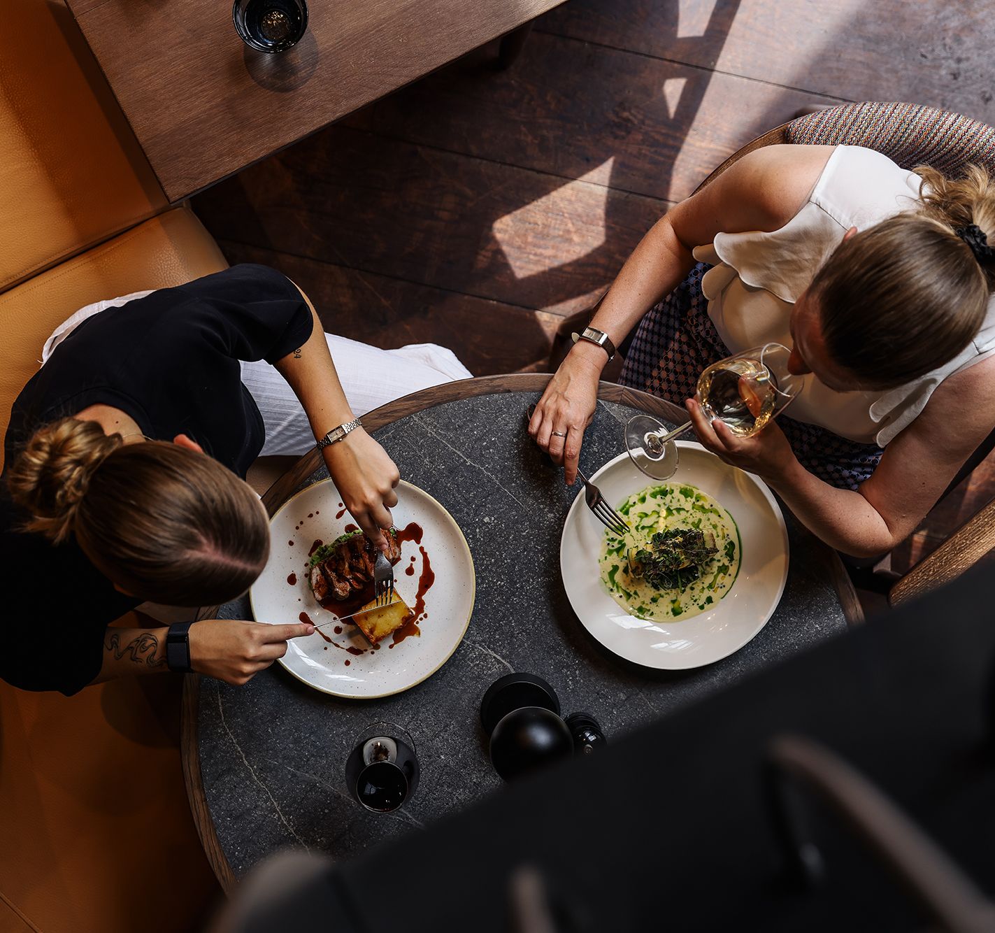 Two women dining at a restaurant table, viewed from above, enjoying plated gourmet meals and drinks.