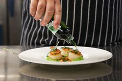 Chef garnishing seared scallops on a white plate with green herb oil.