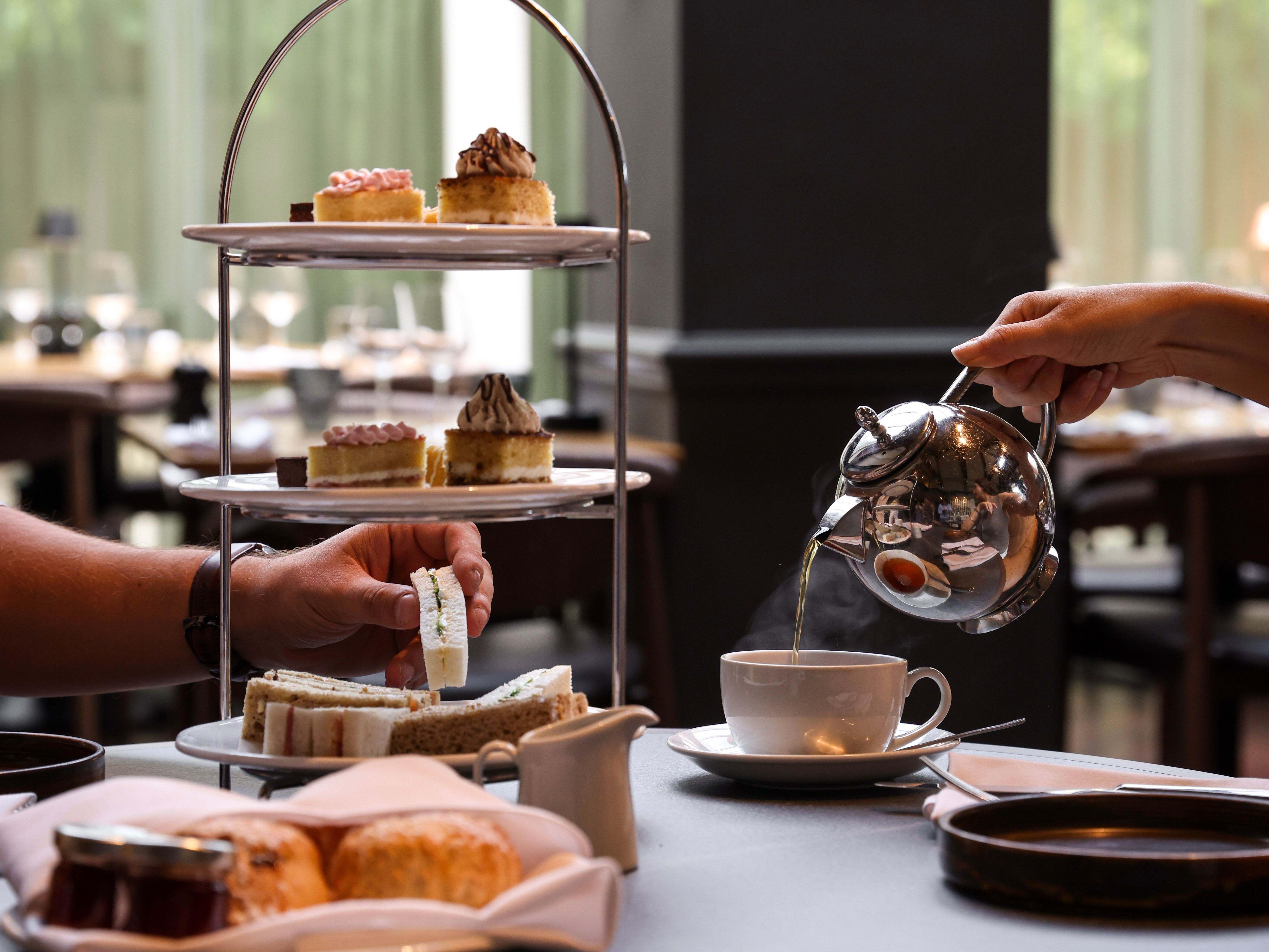 Afternoon tea with a tiered tray of cakes and sandwiches, a person pouring tea into a cup.