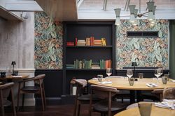 A stylish restaurant interior with round wooden tables, chairs, and a book-filled wall shelf against botanical wallpaper.