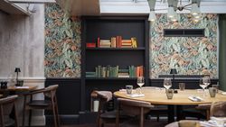 A stylish restaurant interior with round wooden tables, chairs, and a book-filled wall shelf against botanical wallpaper.