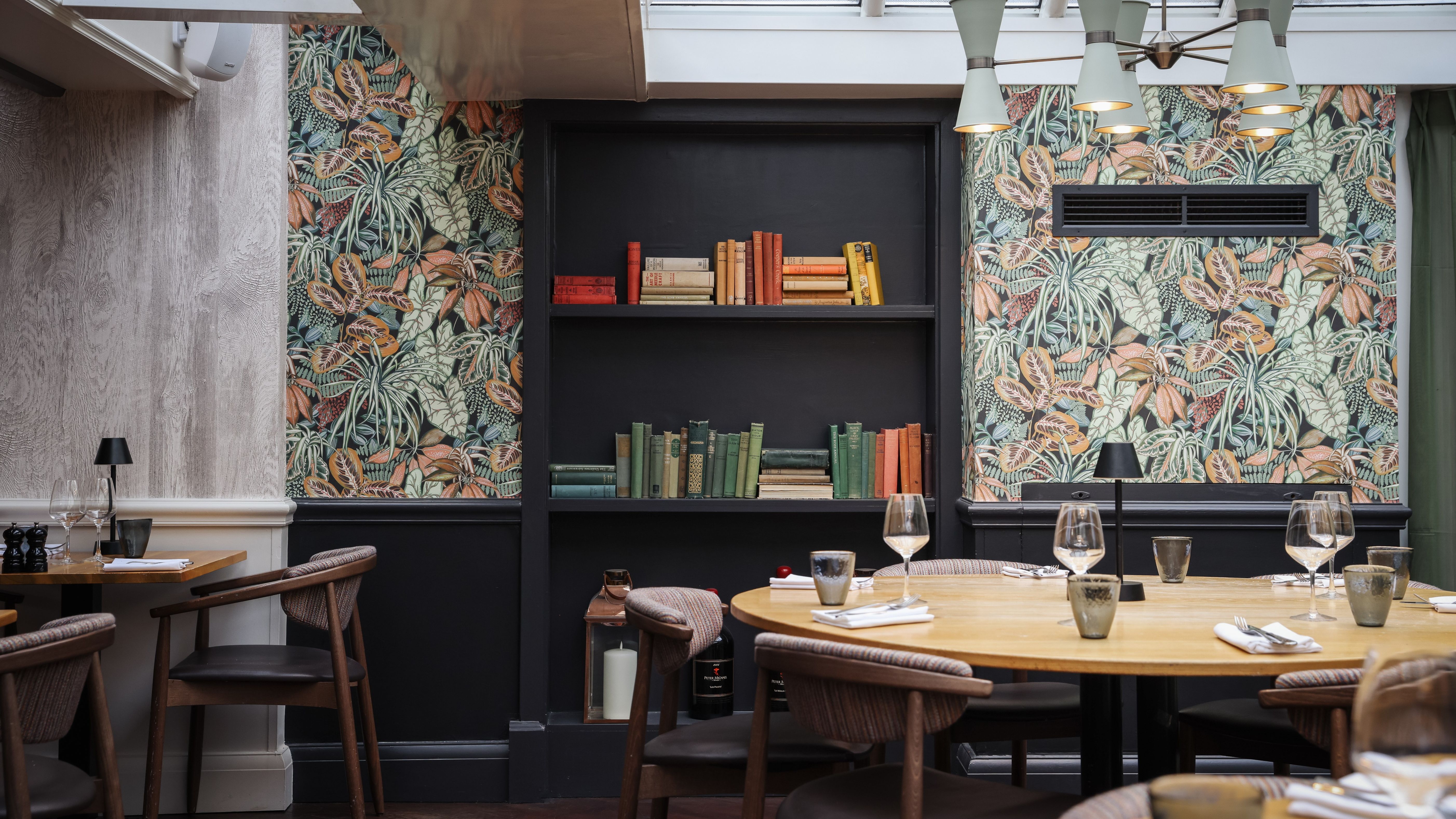 A stylish restaurant interior with round wooden tables, chairs, and a book-filled wall shelf against botanical wallpaper.
