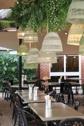 Outdoor seating area of a restaurant with greenery, hanging woven lamps, and tables set with metallic containers.