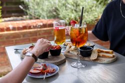 Outdoor table with drinks and assorted appetizers, including bread, cold cuts, and olives, enjoyed by two people.