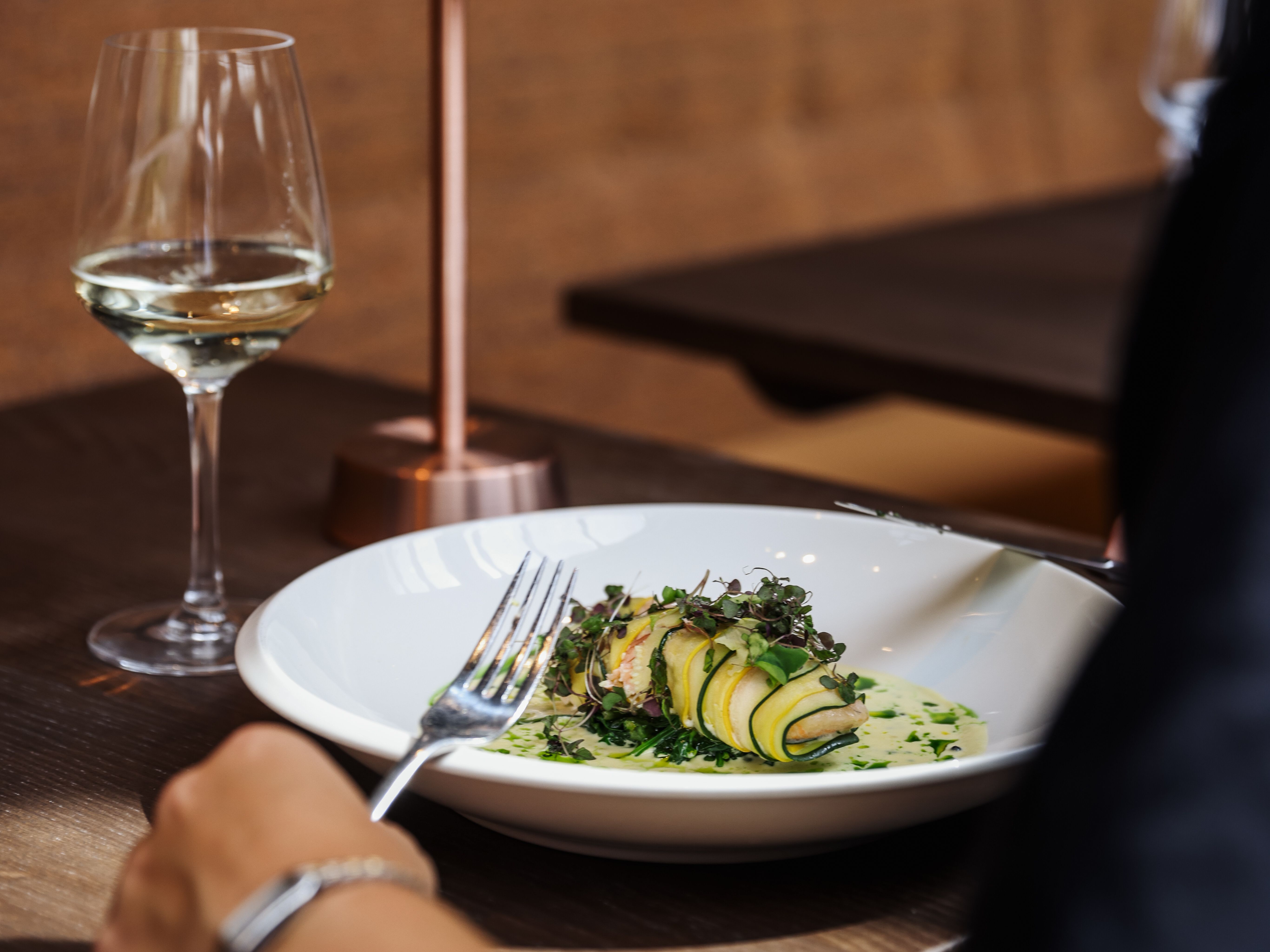 A person dining at a restaurant with a plate of elegantly plated food, a glass of white wine, and a small copper table lamp