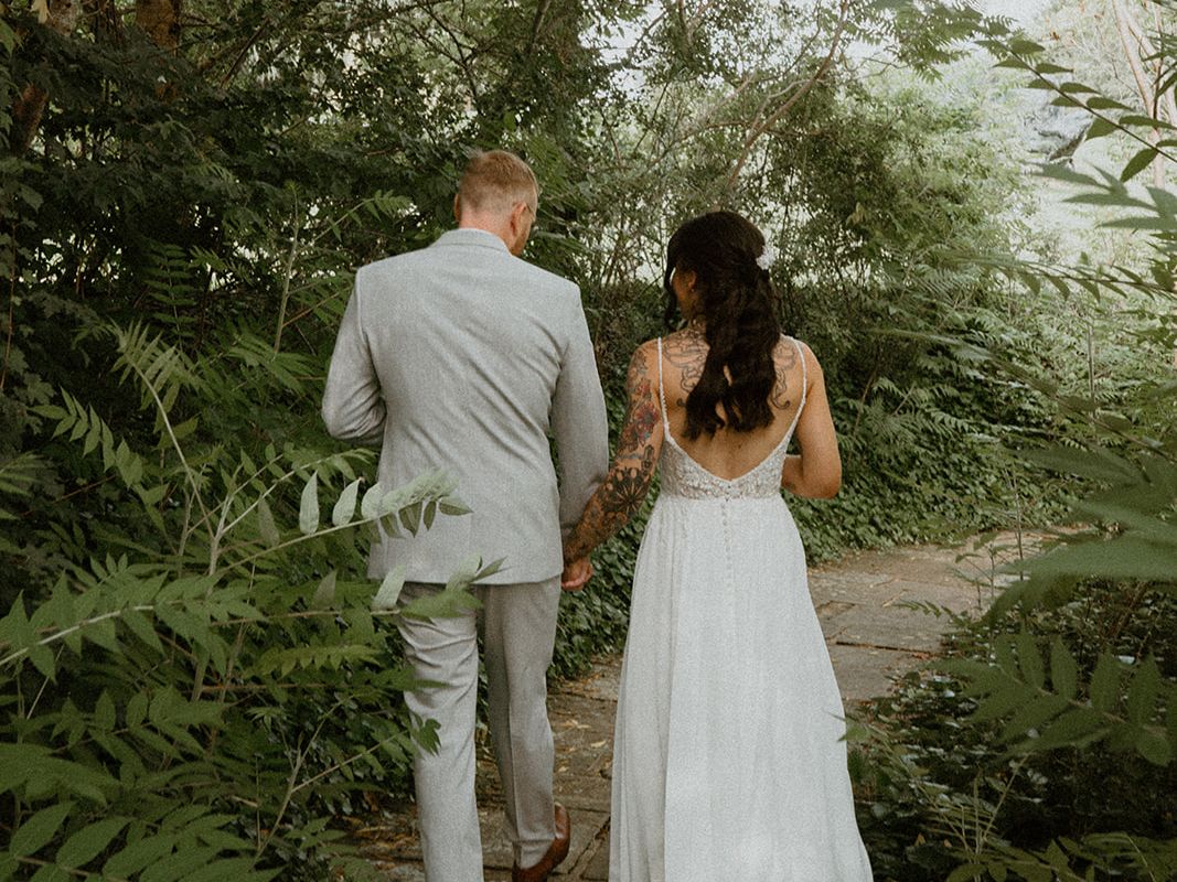 Bride and groom holding hands walking along a stone path surrounded by lush greenery.