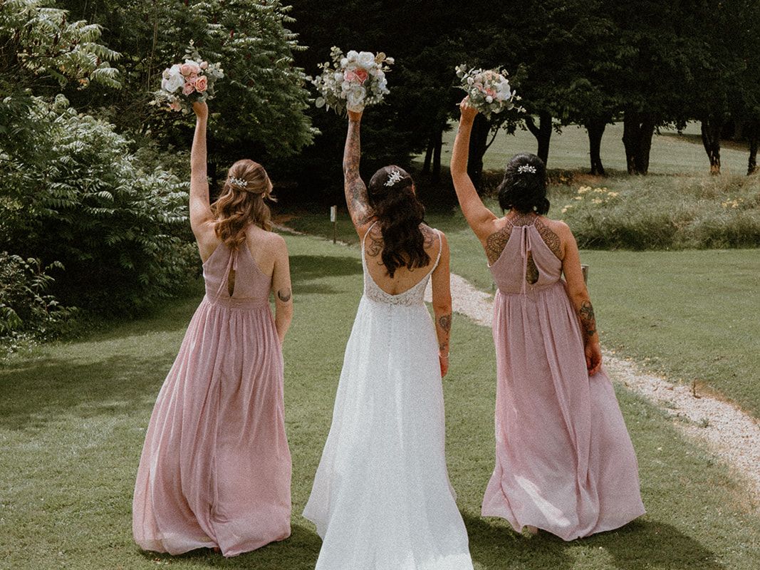 Bride and bridesmaids with bouquets held high walking away on a grassy path outdoors