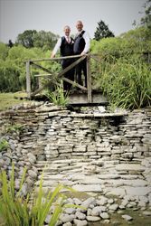 Two men in formal wear standing on a wooden bridge over a stone-lined dry creek in a lush garden setting.