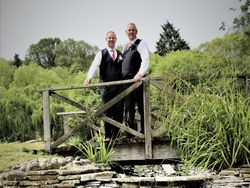 Two men in formal wear standing on a wooden bridge over a stone-lined dry creek in a lush garden setting.