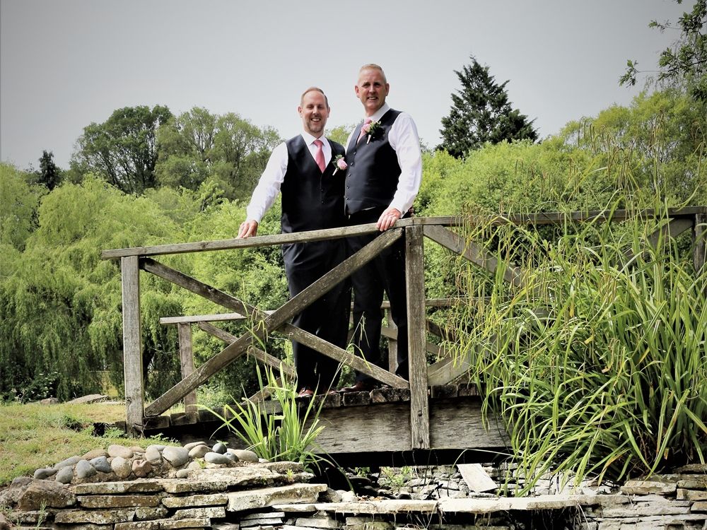 Two men in formal wear standing on a wooden bridge over a stone-lined dry creek in a lush garden setting.