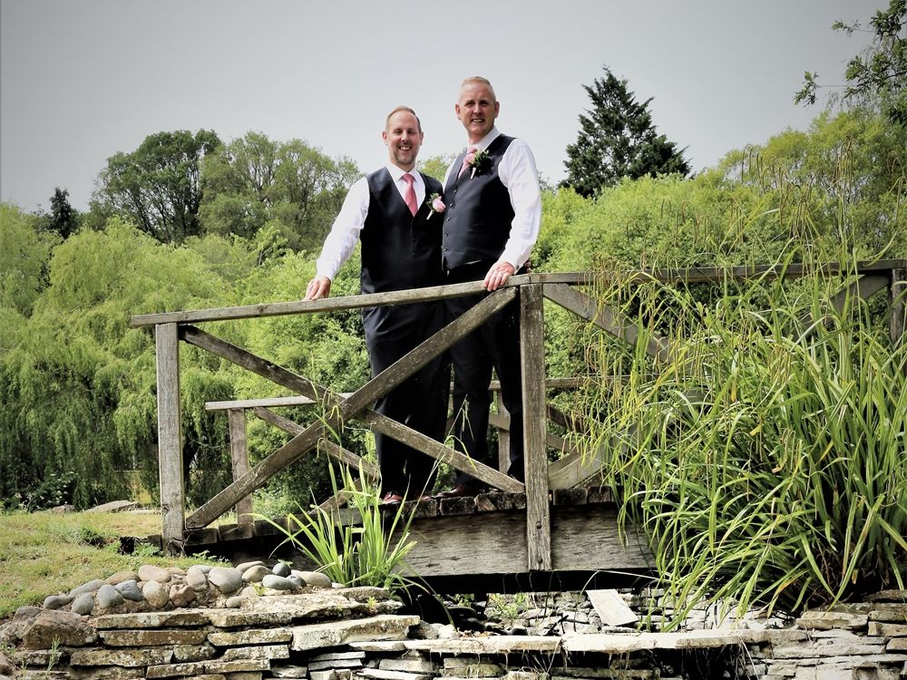 Two men in formal wear standing on a wooden bridge over a stone-lined dry creek in a lush garden setting.