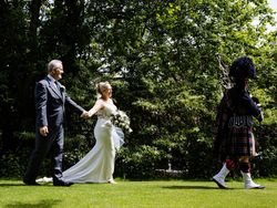 Bride and groom walking hand in hand outdoors, following a bagpiper in traditional Scottish attire.