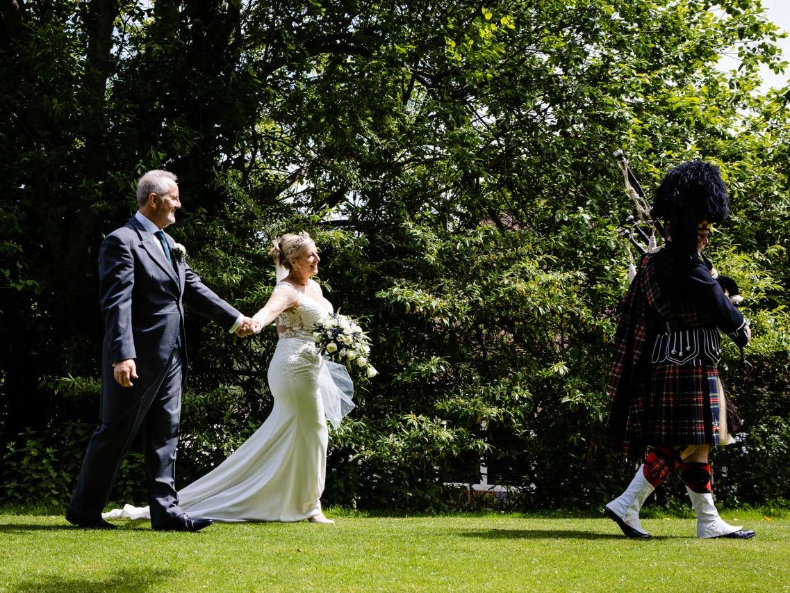 Bride and groom walking hand in hand outdoors, following a bagpiper in traditional Scottish attire.