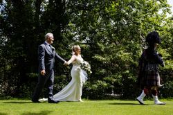 Bride and groom walking hand in hand outdoors, following a bagpiper in traditional Scottish attire.