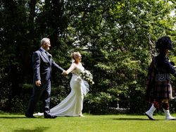 Bride and groom walking hand in hand outdoors, following a bagpiper in traditional Scottish attire.