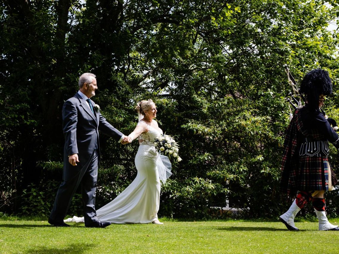 Bride and groom walking hand in hand outdoors, following a bagpiper in traditional Scottish attire.