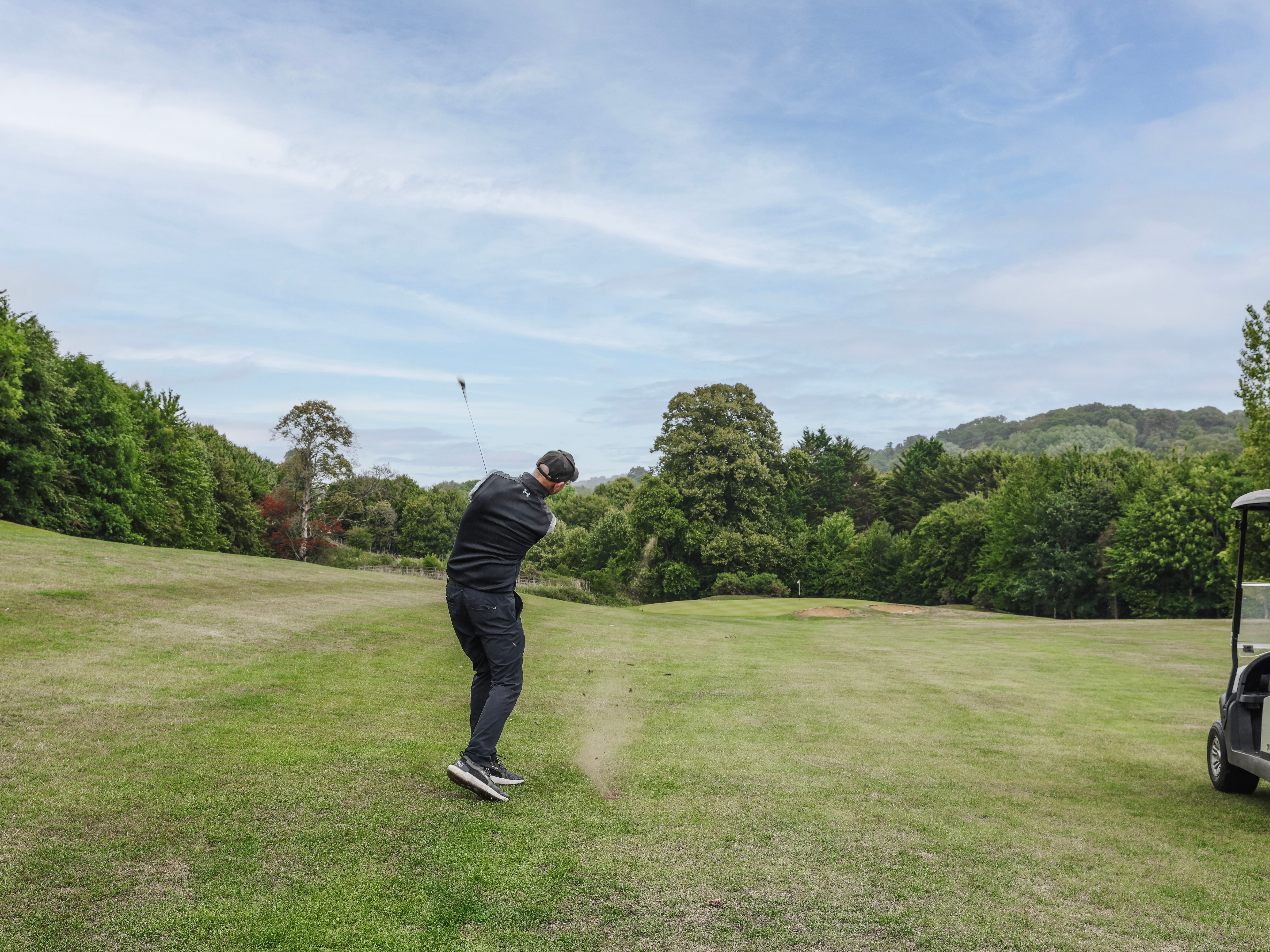 Golfer swinging a club on a lush, green golf course with a golf cart nearby