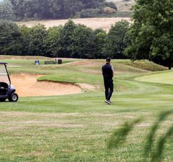 Man standing on a golf course near two golf carts and a sand bunker, surrounded by trees and rolling hills.