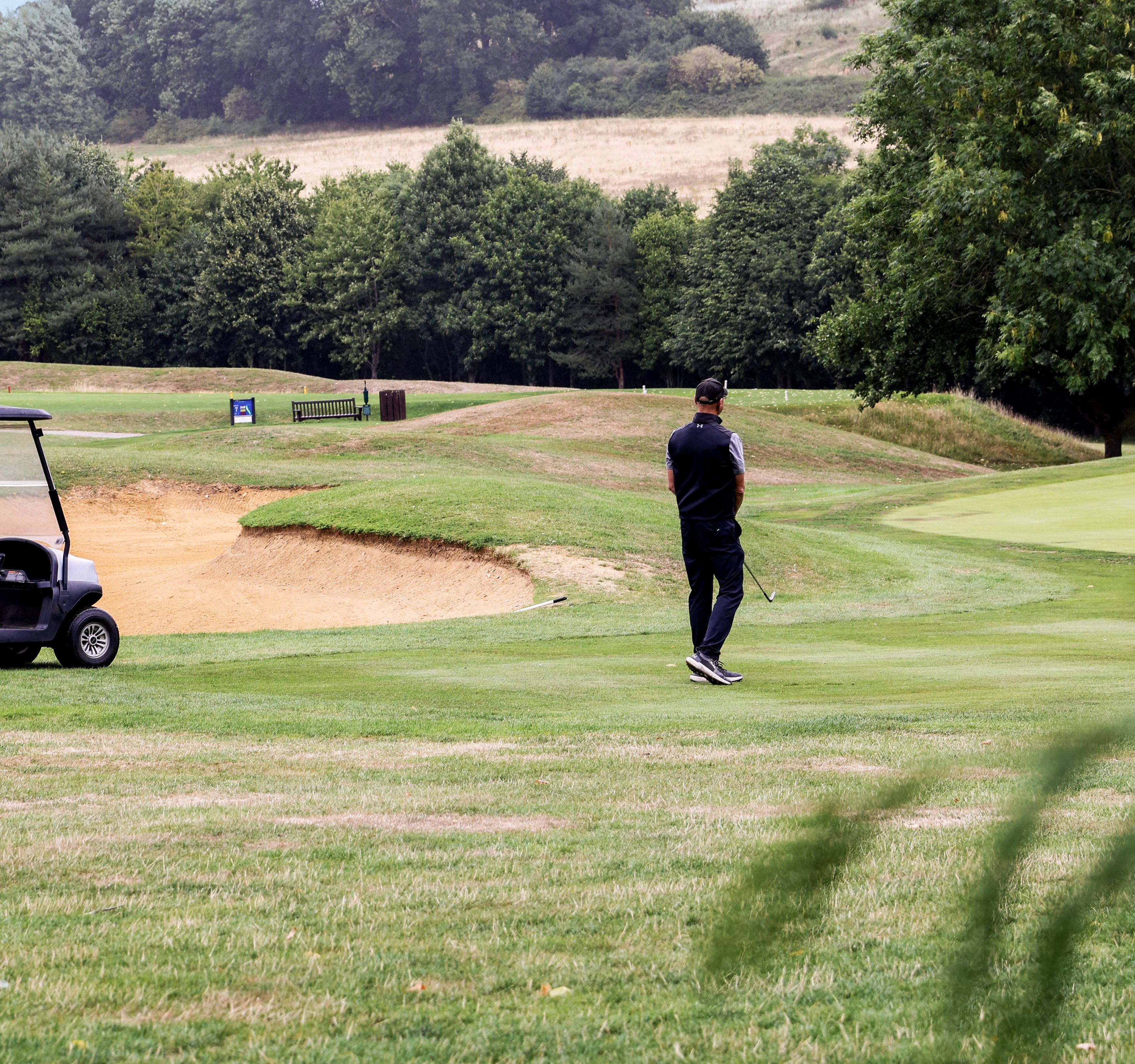 Man standing on a golf course near two golf carts and a sand bunker, surrounded by trees and rolling hills.