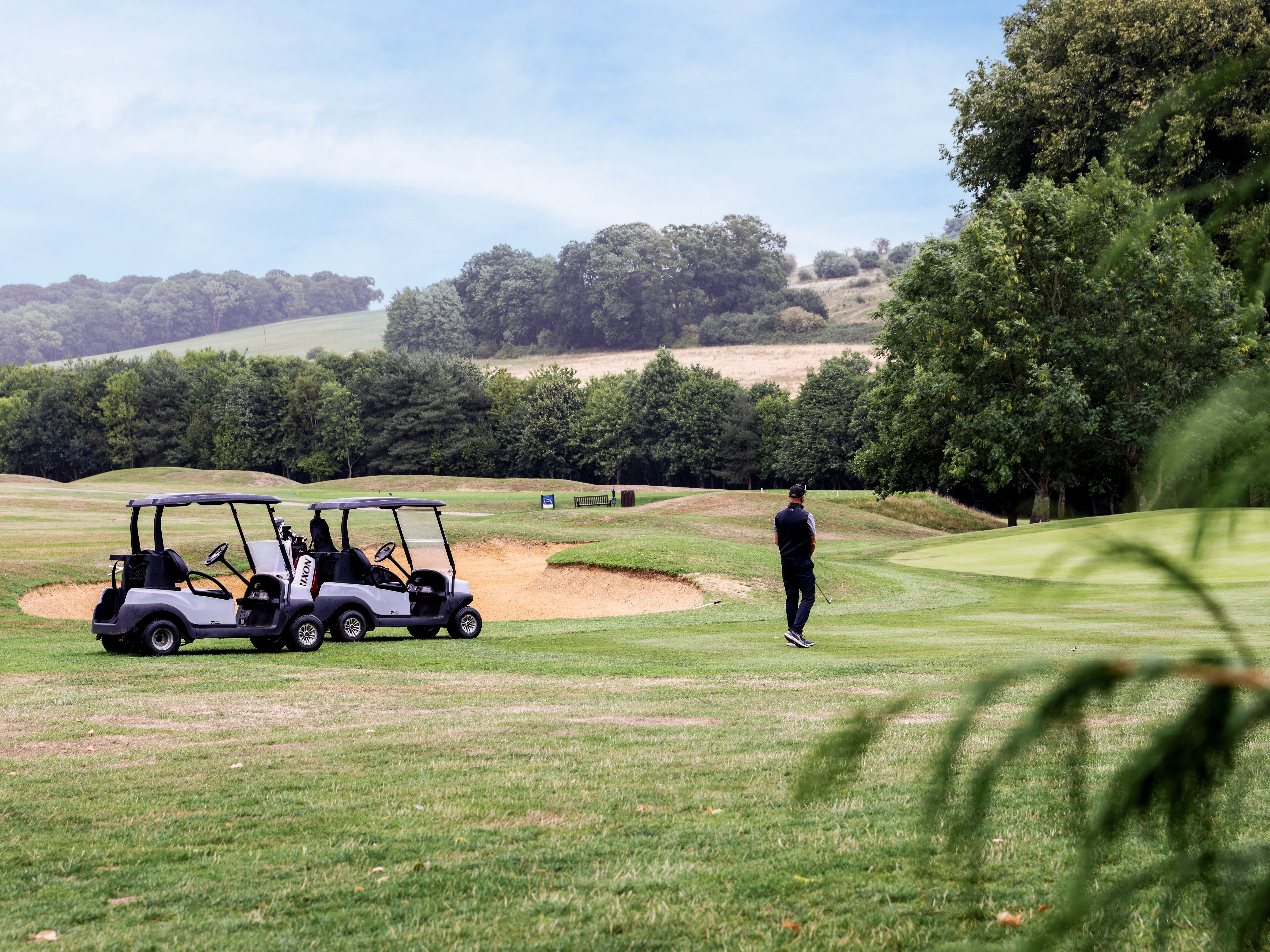 Man standing on a golf course near two golf carts and a sand bunker, surrounded by trees and rolling hills.