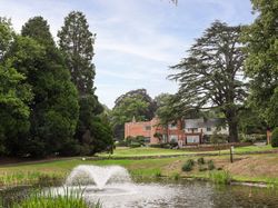 A large brick house surrounded by trees, with a pond and fountain in the foreground.