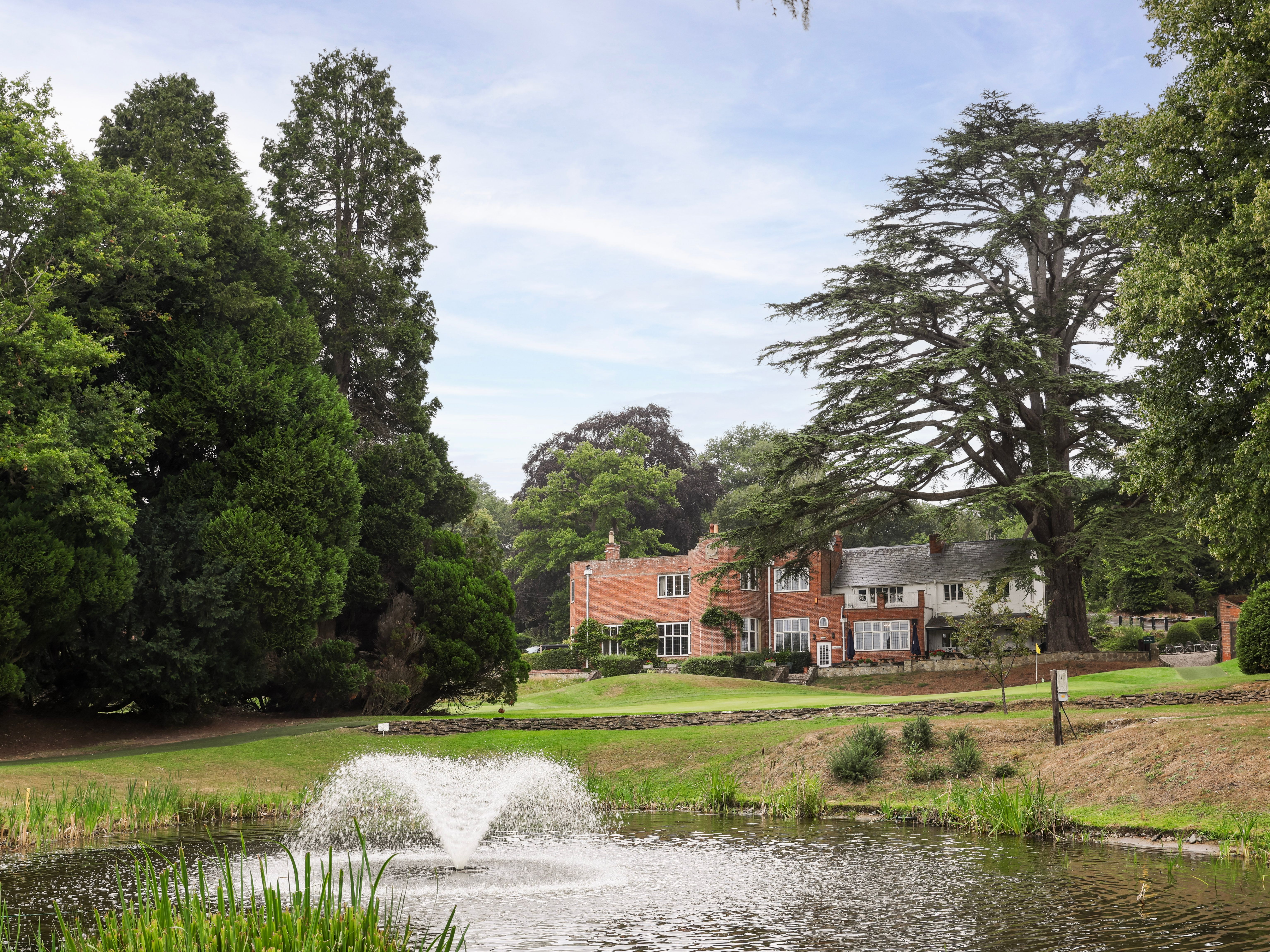 A large brick house surrounded by trees, with a pond and fountain in the foreground.