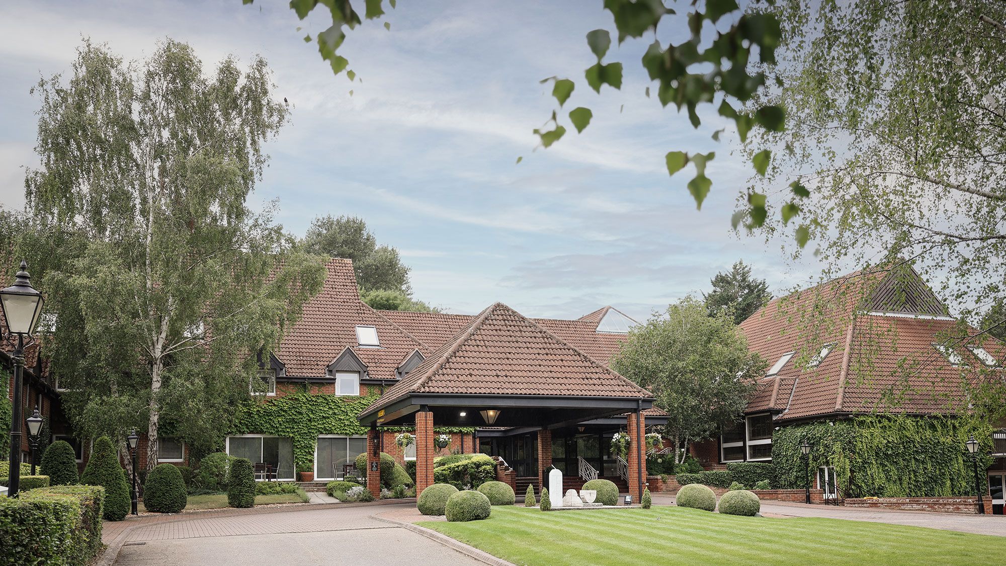 A large brick building with red tiled roof surrounded by lush greenery and manicured lawns.