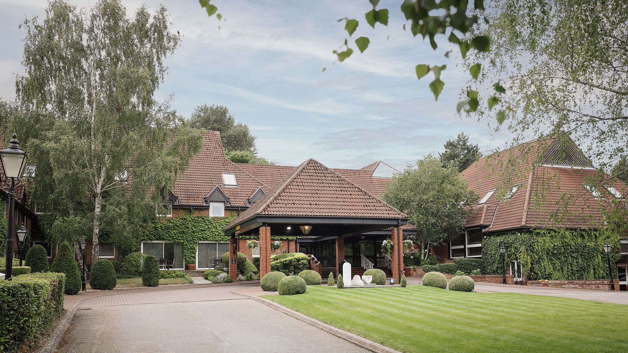 A large brick building with red tiled roof surrounded by lush greenery and manicured lawns.