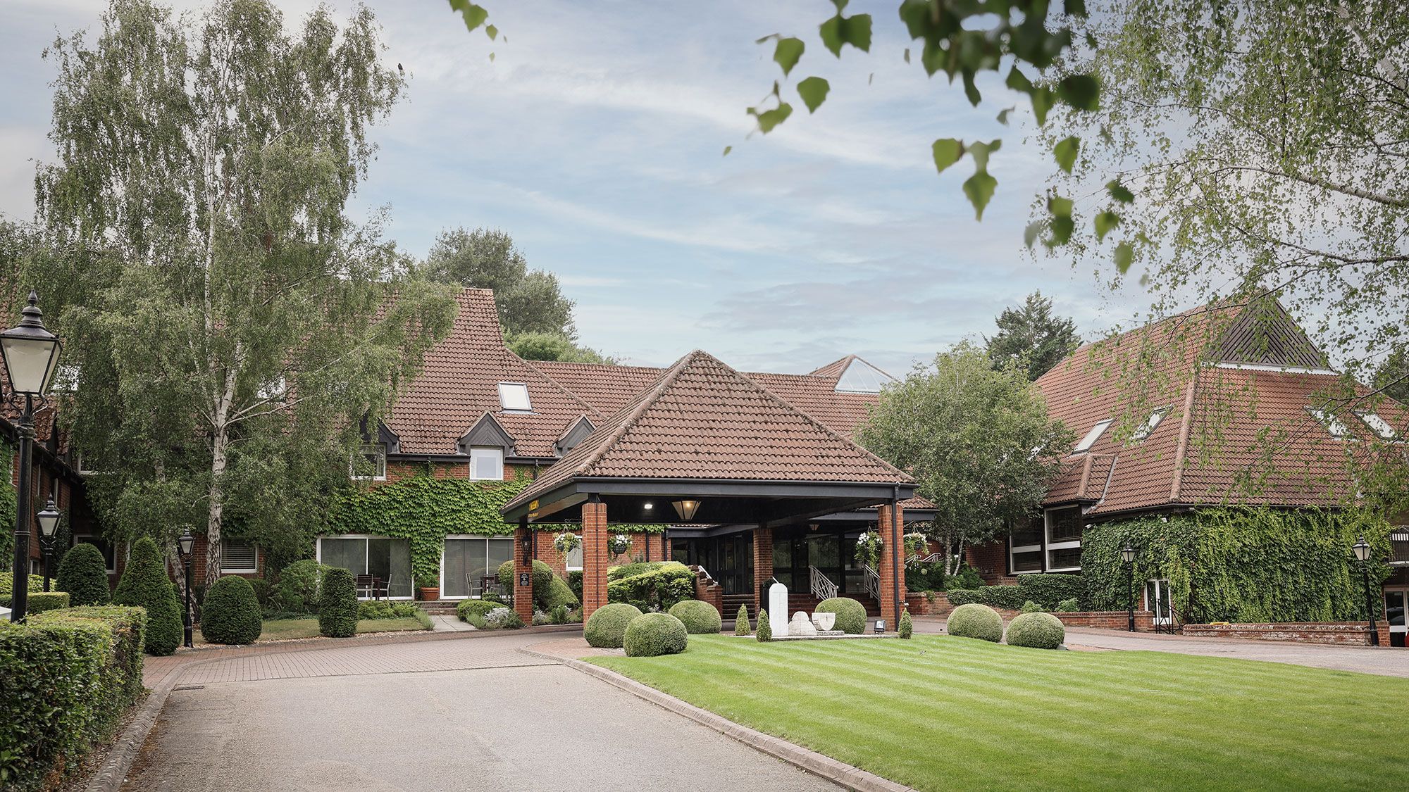 A large brick building with red tiled roof surrounded by lush greenery and manicured lawns.