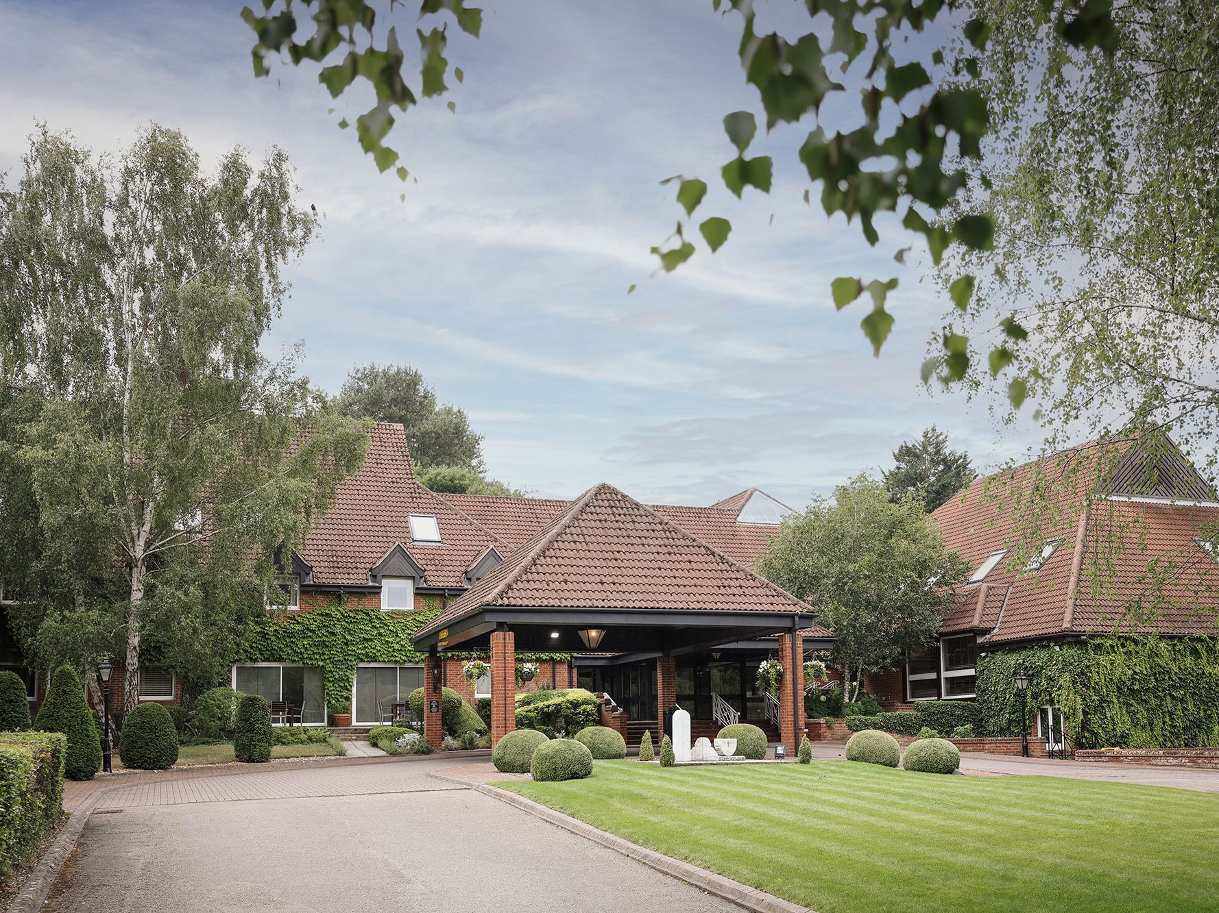 A large brick building with red tiled roof surrounded by lush greenery and manicured lawns.
