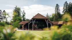A large rustic barn with a red tiled roof and wooden support beams, surrounded by green grass and trees.