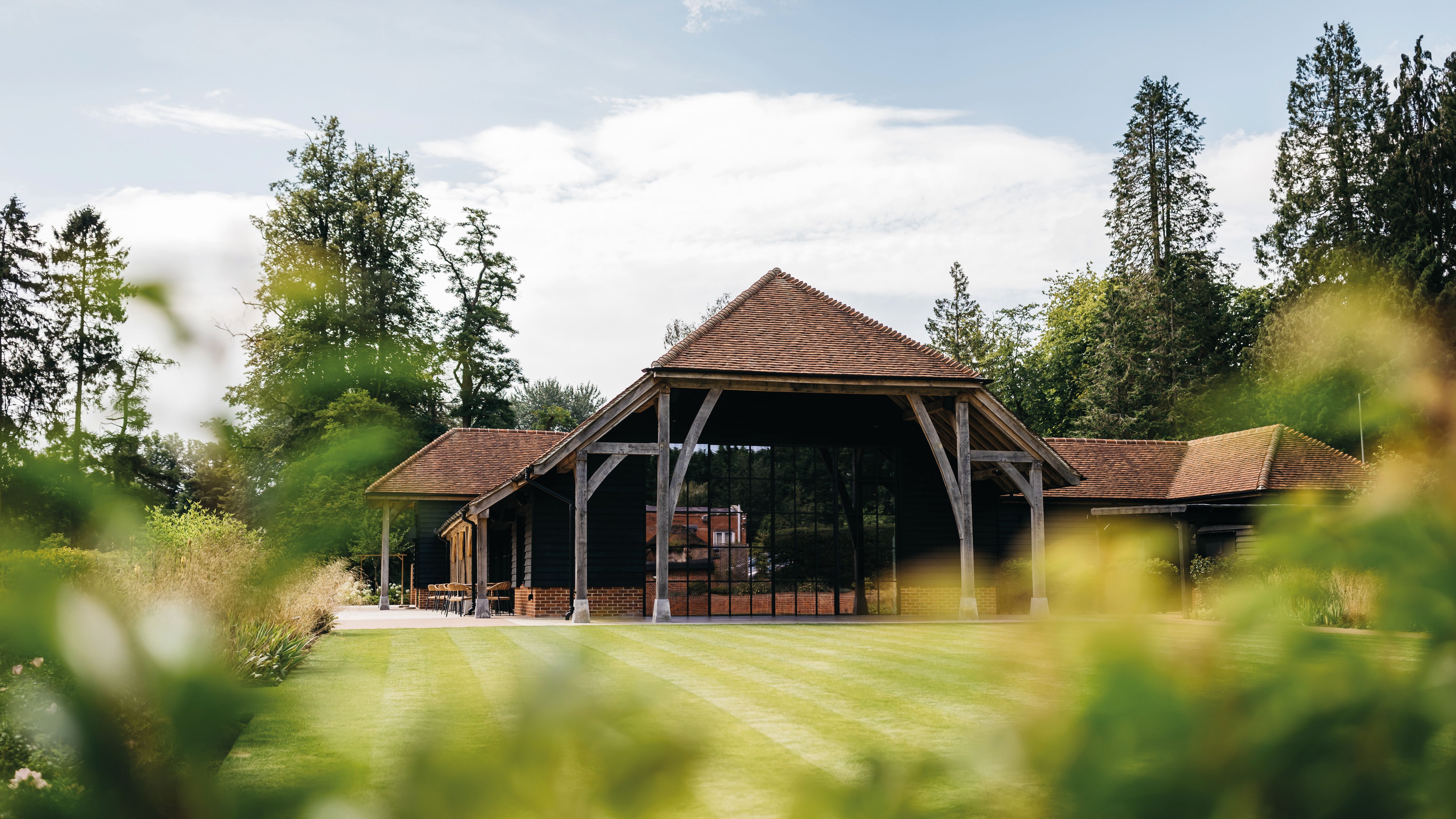 A large rustic barn with a red tiled roof and wooden support beams, surrounded by green grass and trees.
