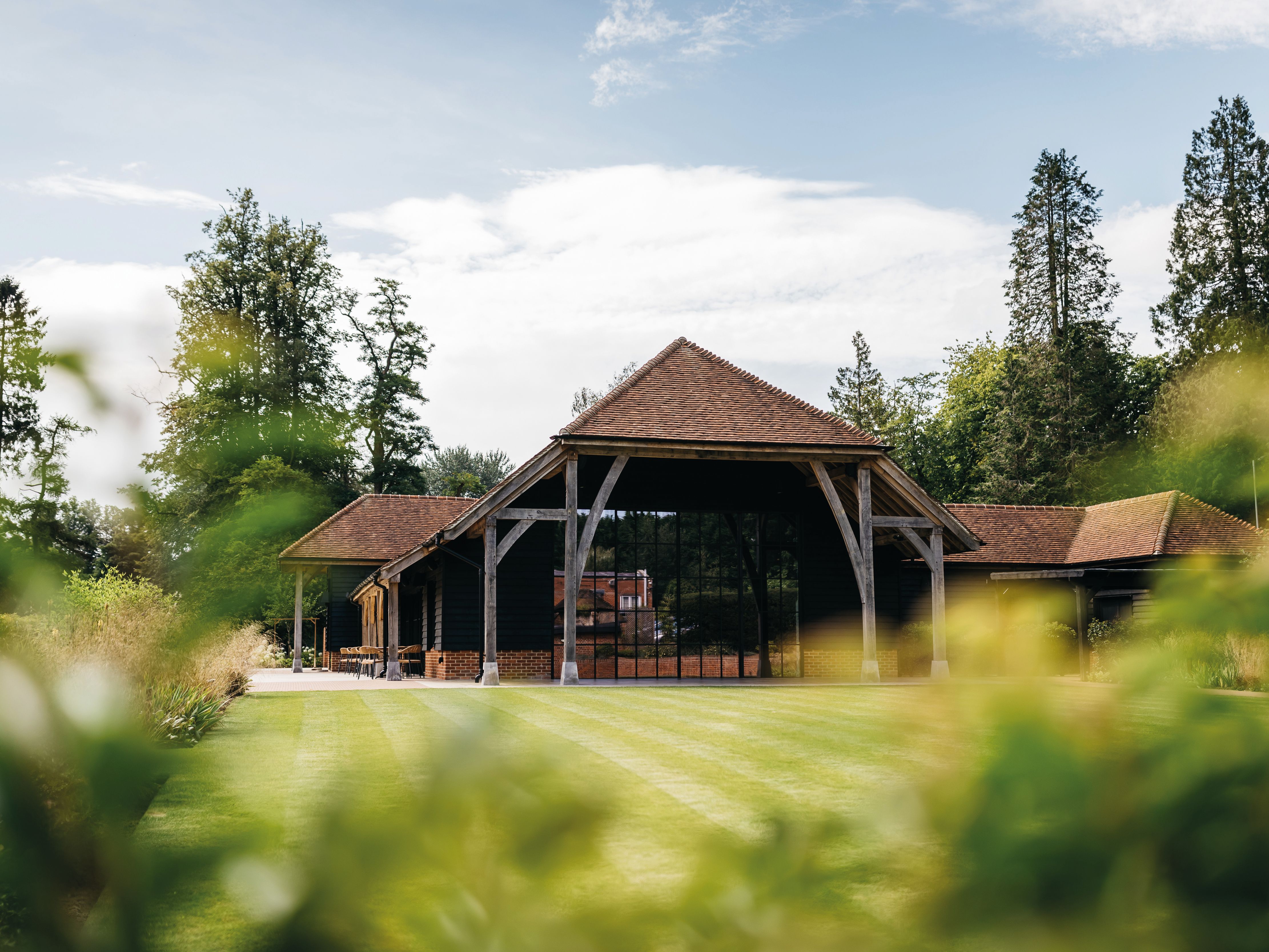 A large rustic barn with a red tiled roof and wooden support beams, surrounded by green grass and trees.