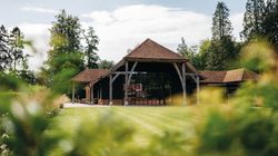 A large rustic barn with a red tiled roof and wooden support beams, surrounded by green grass and trees.