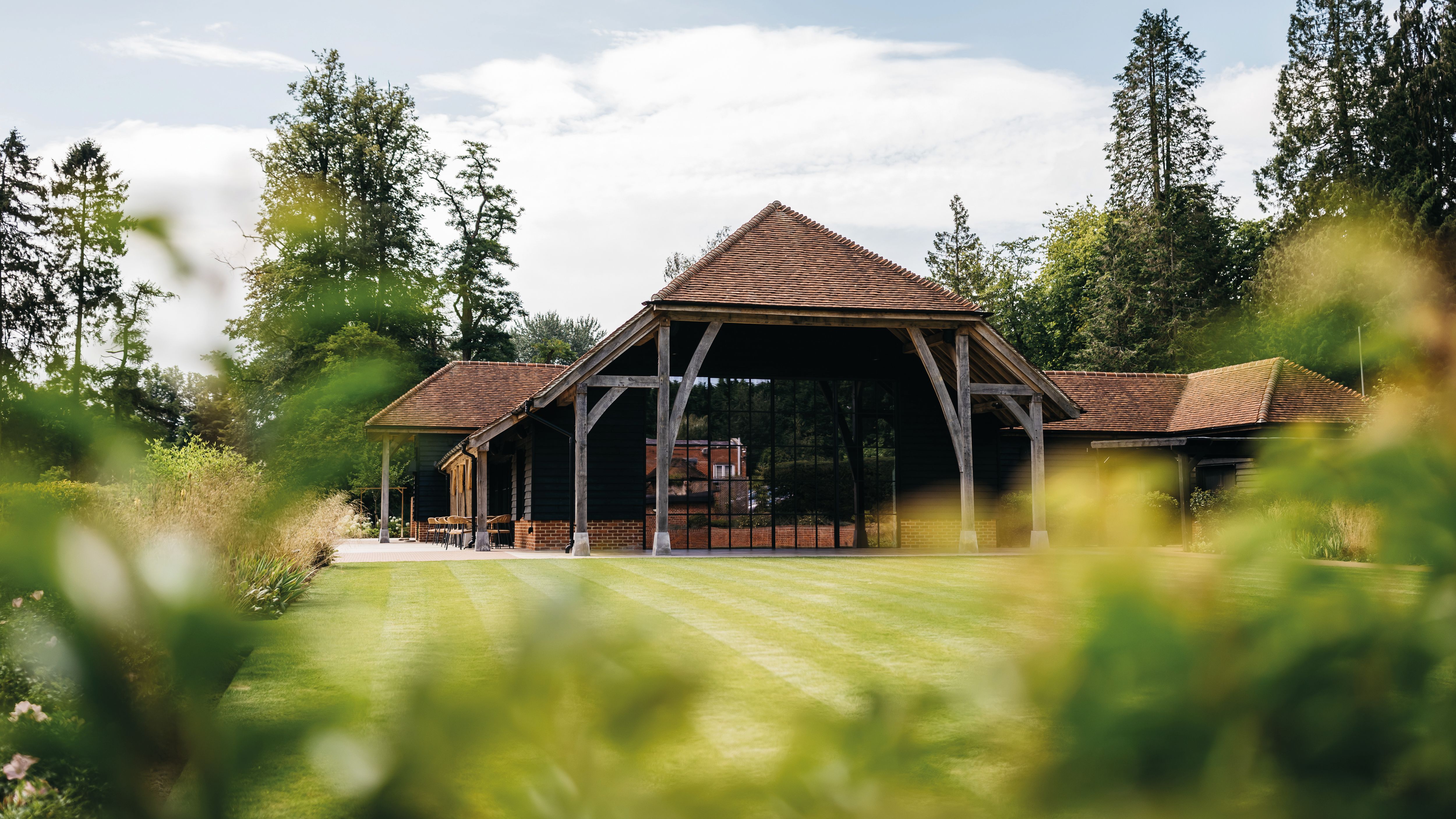 A large rustic barn with a red tiled roof and wooden support beams, surrounded by green grass and trees.