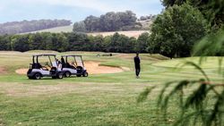 Person standing near two golf carts on a grassy golf course surrounded by trees, with a sand bunker and hills in the background.