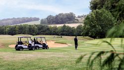 Person standing near two golf carts on a grassy golf course surrounded by trees, with a sand bunker and hills in the background.