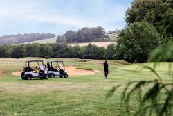 Person standing near two golf carts on a grassy golf course surrounded by trees, with a sand bunker and hills in the background.