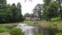 Pond with water fountain in front of a large brick house surrounded by trees and green lawn.