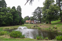 Pond with water fountain in front of a large brick house surrounded by trees and green lawn.