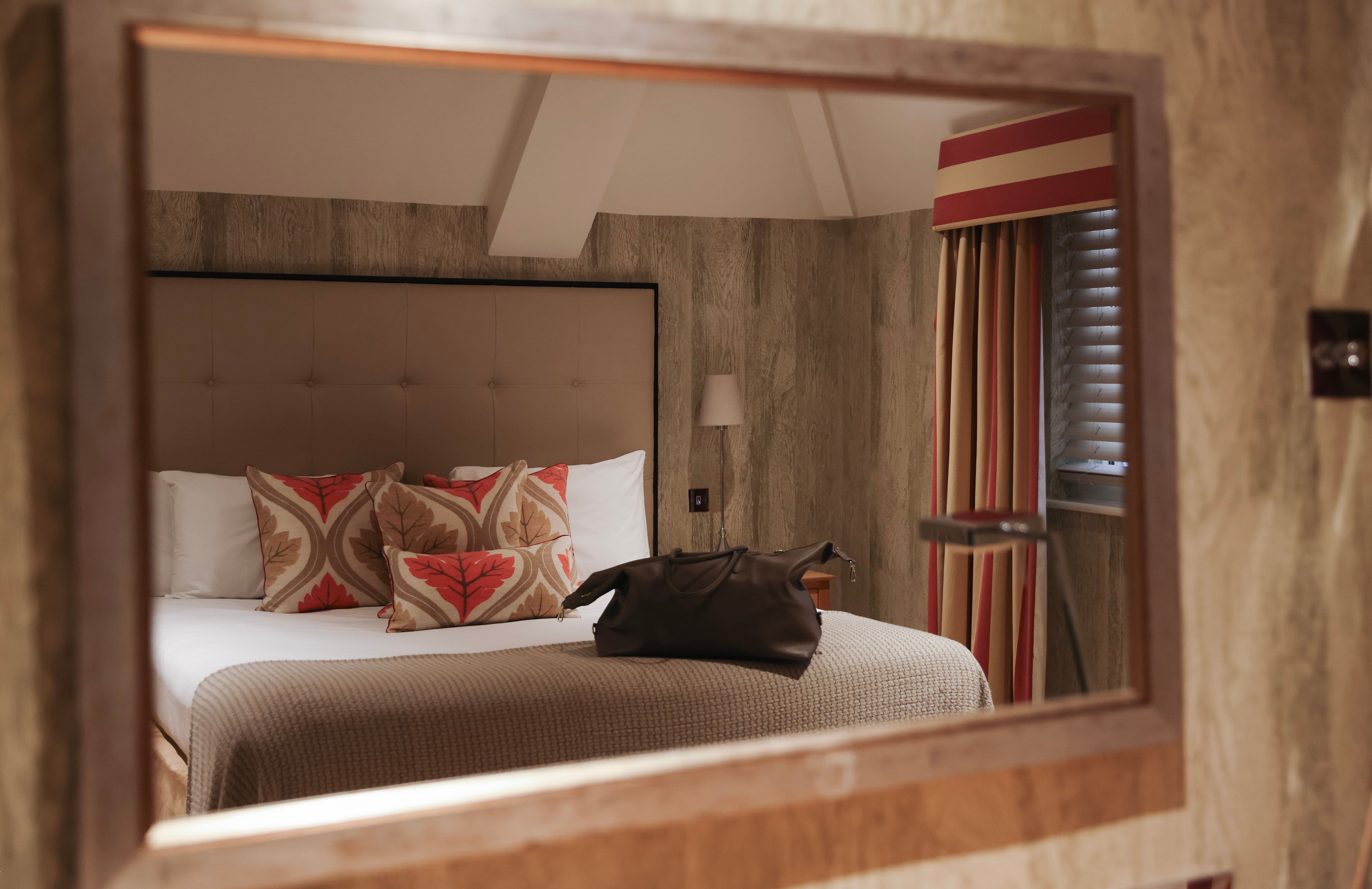 Reflection of a cozy bedroom with patterned pillows and a bag on the bed seen in a wall mirror.