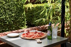 Pepperoni pizza on a wooden board, Peroni beer, and a glass of sparkling water with lemon on an outdoor table surrounded by greenery.
