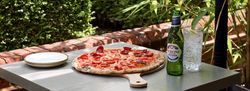 Pepperoni pizza on a wooden board, Peroni beer, and a glass of sparkling water with lemon on an outdoor table surrounded by greenery.