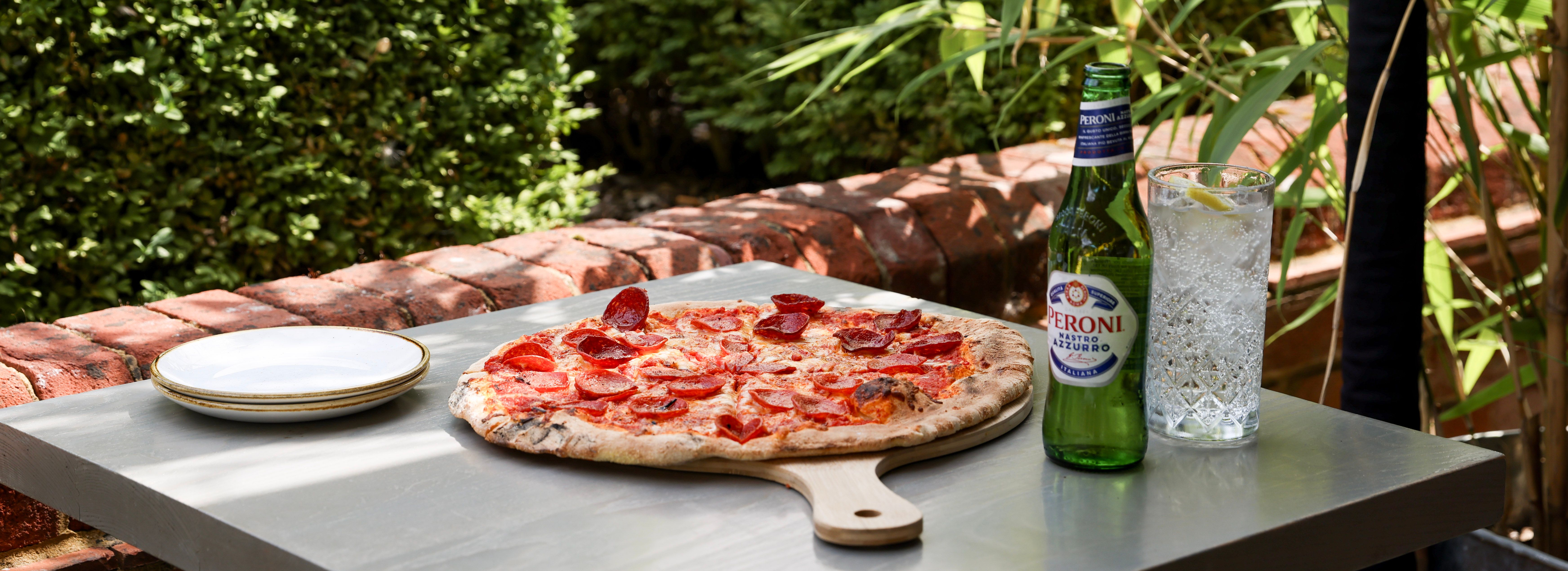 Pepperoni pizza on a wooden board, Peroni beer, and a glass of sparkling water with lemon on an outdoor table surrounded by greenery.