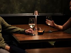 Two people sitting at a dimly lit table with wine glasses and a small dish of snacks, beneath a modern lamp.