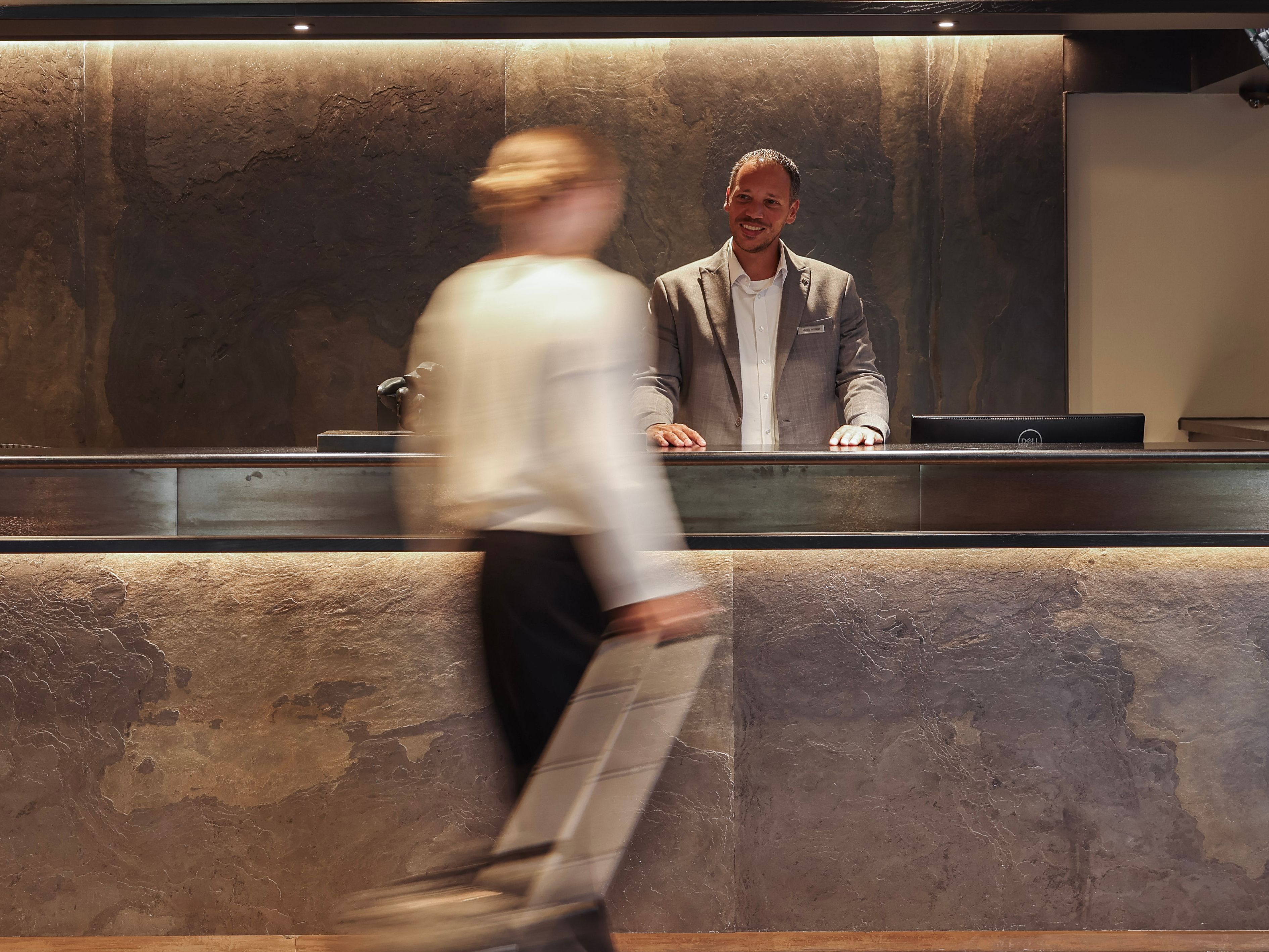 Hotel lobby reception desk with a receptionist standing behind the counter and a guest walking by with luggage.