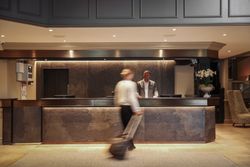Hotel lobby reception desk with a receptionist standing behind the counter and a guest walking by with luggage.