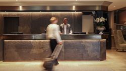 Hotel lobby reception desk with a receptionist standing behind the counter and a guest walking by with luggage.